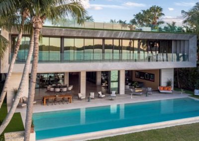 Modern two-story house with glass walls, pool, outdoor seating, palm trees, and a water view at sunset.