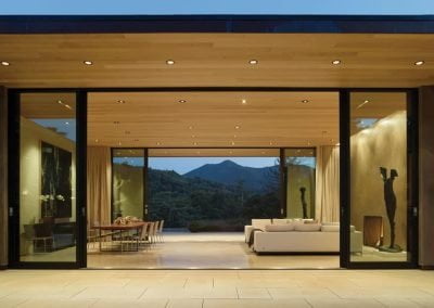 Modern open living room with large glass walls, wood ceiling, mountain view, and sculptures inside.