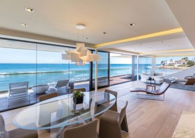 Modern beachfront living room with glass walls, ocean view, dining area, lounge chairs, and natural sunlight.