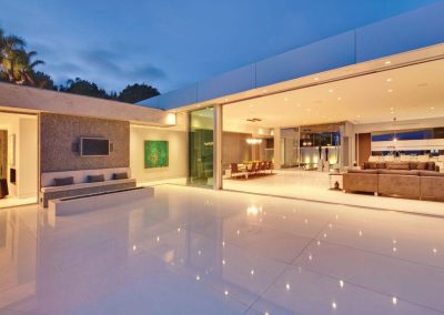 Modern luxury home with large open glass walls, spacious patio, and warm interior lighting at dusk.