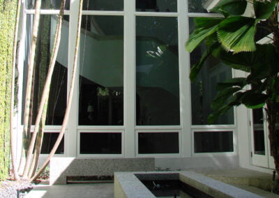Tall windows reflect greenery; modern patio features stone bench, lush plants, and geometric water feature in sunlight.