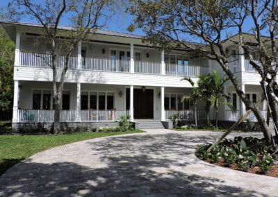 Two-story white house with wraparound porch, large windows, and a circular driveway, surrounded by trees and greenery.