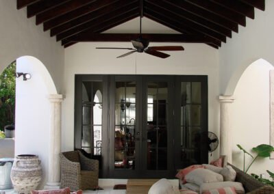 Open covered patio with wicker furniture, arched pillars, ceiling fan, and double glass doors leading inside.