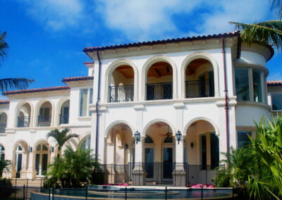 Large, white Mediterranean-style mansion with arches, balconies, palm trees, and a pool under a blue sky.