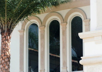 Three arched windows with beige trim reflect palm trees, next to a stone column and a palm tree trunk in front.