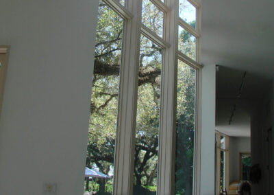 Tall windows in a white room reveal trees and greenery outside, creating a bright, airy atmosphere with natural light.