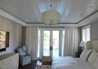 Bright bedroom with white bed, armchair, TV, dresser, and large windows with white curtains facing a balcony.