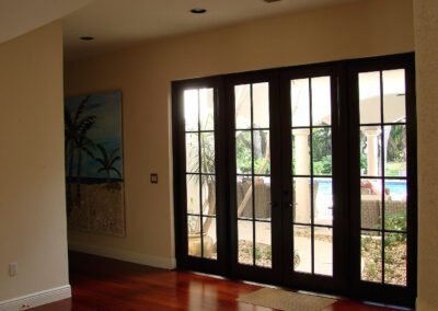French doors open to a patio with a pool, tropical plants, wicker furniture, and a painting of palm trees inside.