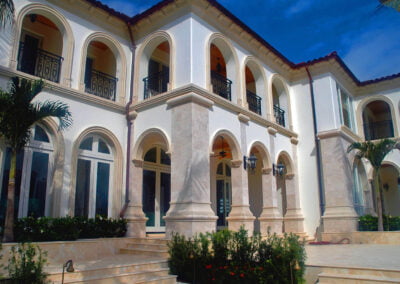 Large Mediterranean-style mansion with white walls, arched windows, balconies, palm trees, and blue sky.
