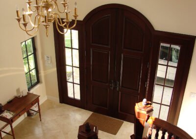 Large dark wooden double doors with glass panels, chandelier, tiled floor, table, and staircase in a bright entryway.