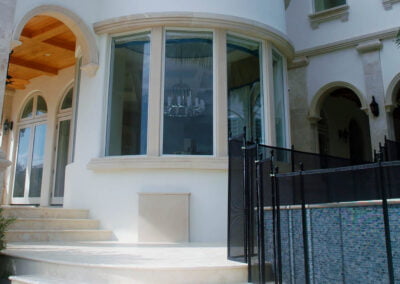 White house exterior with large curved windows, arched doorway, stone steps, and a black pool safety fence.