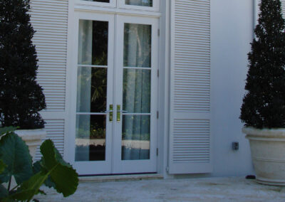 White French doors with glass panes, flanked by tall potted topiary trees and white louvered shutters.