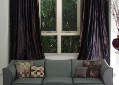 Gray sofa with patterned pillows sits in front of a window with dark curtains and greenery visible outside.
