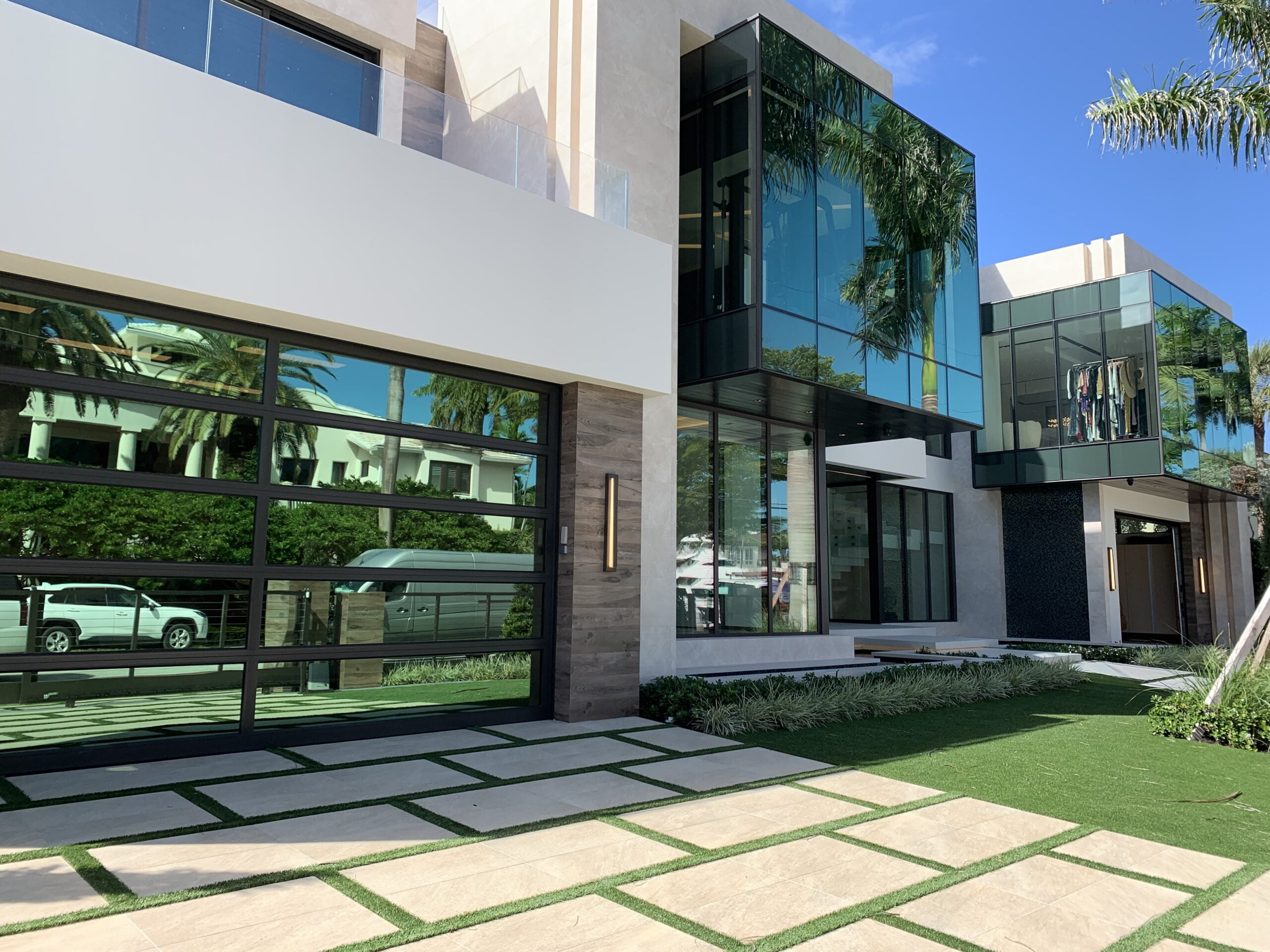 Modern house with large glass windows, a glass garage door, palm trees, and a neatly paved driveway under clear skies.